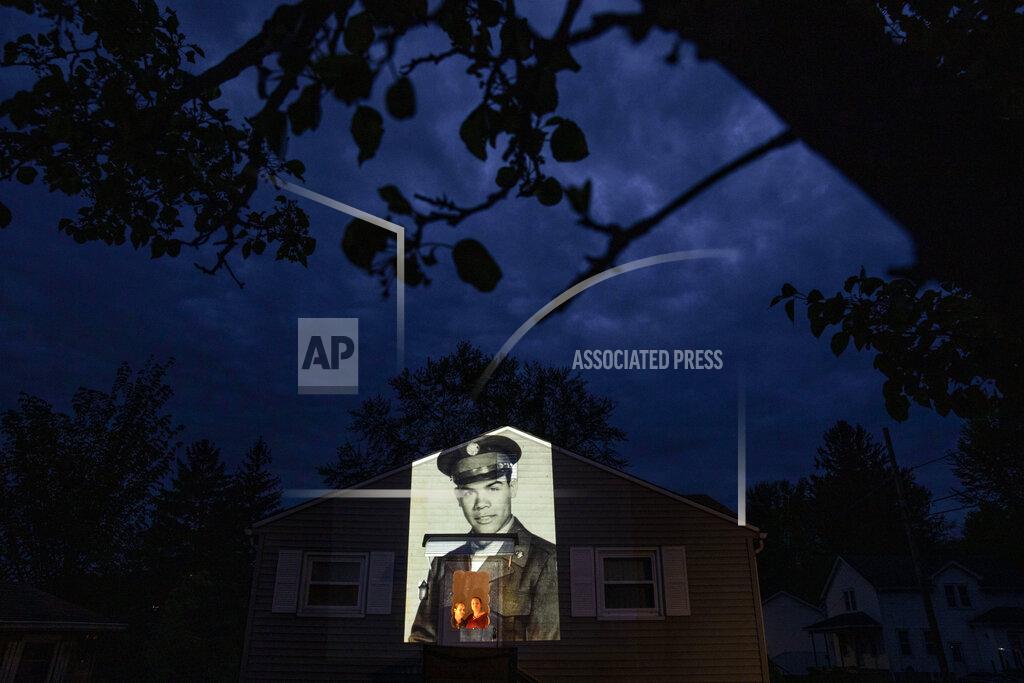 David Goldman An image of veteran Samuel Melendez is projected onto the home of his nieces, Janet Ramirez, right, and Mary Perez, as they look out a doorway in Chicopee, Mass., Sunday, May 17, 2020. Originally of Puerto Rico, Melendez, a U.S. Army Korean War veteran and resident of the Soldier's Home in Holyoke, Mass., died from the COVID-19 virus at the age of 86. Seeking to capture moments of private mourning at a time of global isolation, the photographer used a projector to cast large images of veterans on to the homes as their loved ones are struggling to honor them during a lockdown that has sidelined many funeral traditions. (AP Photo/David Goldman)