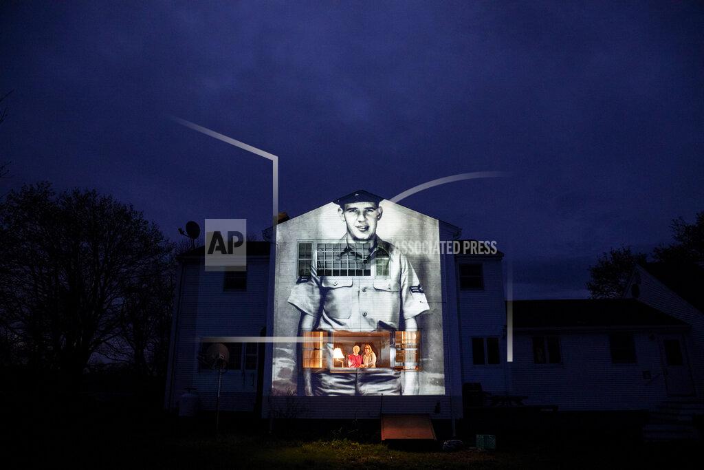 David Goldman An image of veteran Charles Lowell is projected onto the home he shared with his wife, Alice, for 30 years as she stands at left with her daughter, Susan Kenney, in Hardwick, Mass. Saturday, May 2, 2020. Lowell, a U.S. Air Force veteran and resident of the Soldier's Home in Holyoke, Mass., died from the COVID-19 virus at the age of 78. Seeking to capture moments of private mourning at a time of global isolation, the photographer used a projector to cast large images of veterans on to the homes as their loved ones are struggling to honor them during a lockdown that has sidelined many funeral traditions. (AP Photo/David Goldman)
