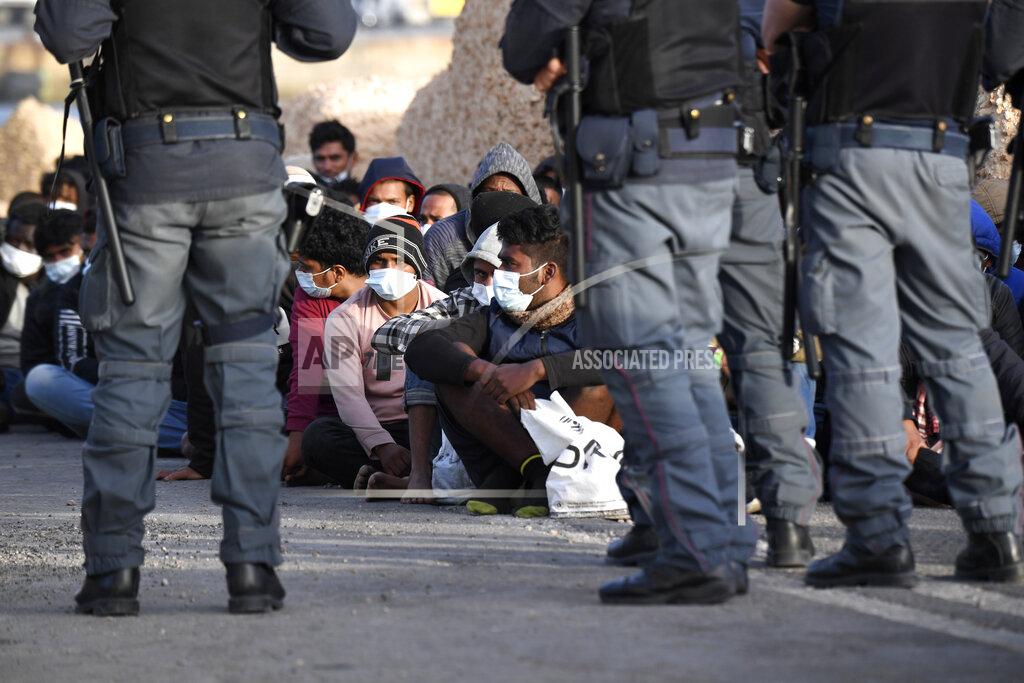Salvatore Cavalli Migrants wearing face masks to curb the spread of COVID-19 sit at a pier as Italian police officers stand by, on the Sicilian island of Lampedusa, southern Italy, Monday, May 10, 2021. Several hundred more migrants reached the tiny Italian island before dawn on Monday, swelling to past 2,100 the number of arrivals in around 24 hours and fueling calls from across the political spectrum for the Italian government to strengthen its migration policies. (AP Photo/Salvatore Cavalli)