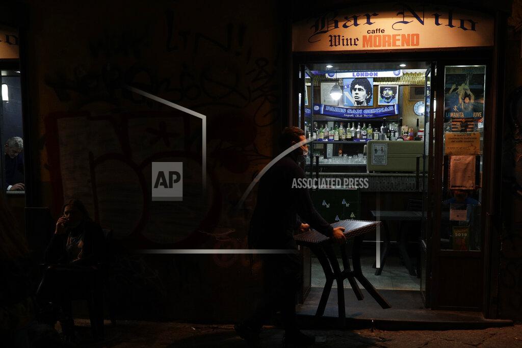 A bartender carries a table inside as he is closing the Nilo Bar in Naples, in the region of Campania, Italy, Friday, Nov. 13, 2020. The regions of Campania and Tuscany were designated red zone on Friday, signaling the dire condition of a hospitals struggling with a surge of new admissions. (AP Photo/Gregorio Borgia)