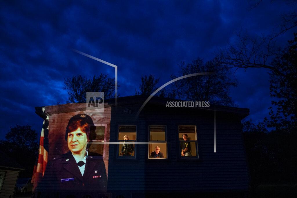 David Goldman An image of veteran Constance "Kandy" Pinard is projected onto the home she grew up in with her sister, Tammy Petrowicz, left, and brothers, Paul, center, and Brian Driscoll in Florence, Mass., Thursday, May 14, 2020. Pinard, a nurse in the U.S. Air Force and resident of the Soldier's Home in Holyoke, Mass., died from the COVID-19 virus at the age of 73. Seeking to capture moments of private mourning at a time of global isolation, the photographer used a projector to cast large images of veterans on to the homes as their loved ones are struggling to honor them during a lockdown that has sidelined many funeral traditions. (AP Photo/David Goldman)