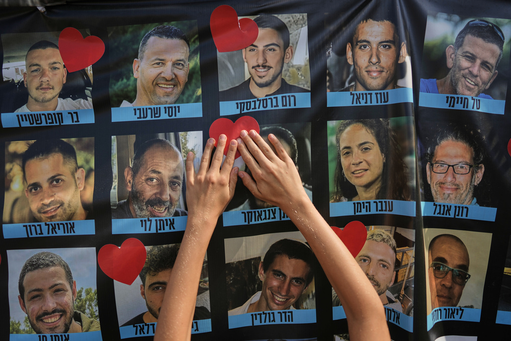 Hands paste a heart-shaped sticker onto a banner with pictures of Israeli hostages during a a gathering at a plaza known as hostages square in Tel Aviv, Israel, Monday, Oct. 13, 2025. (AP Photo/Oded Balilty)