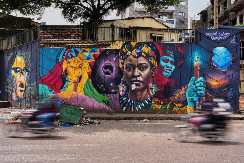 Motorcyclists ride past a mural painted by Graffiti artist Omar Diaw in Conakry, Guinea, Saturday, Sep. 20, 2025. (AP Photo/Misper Apawu)