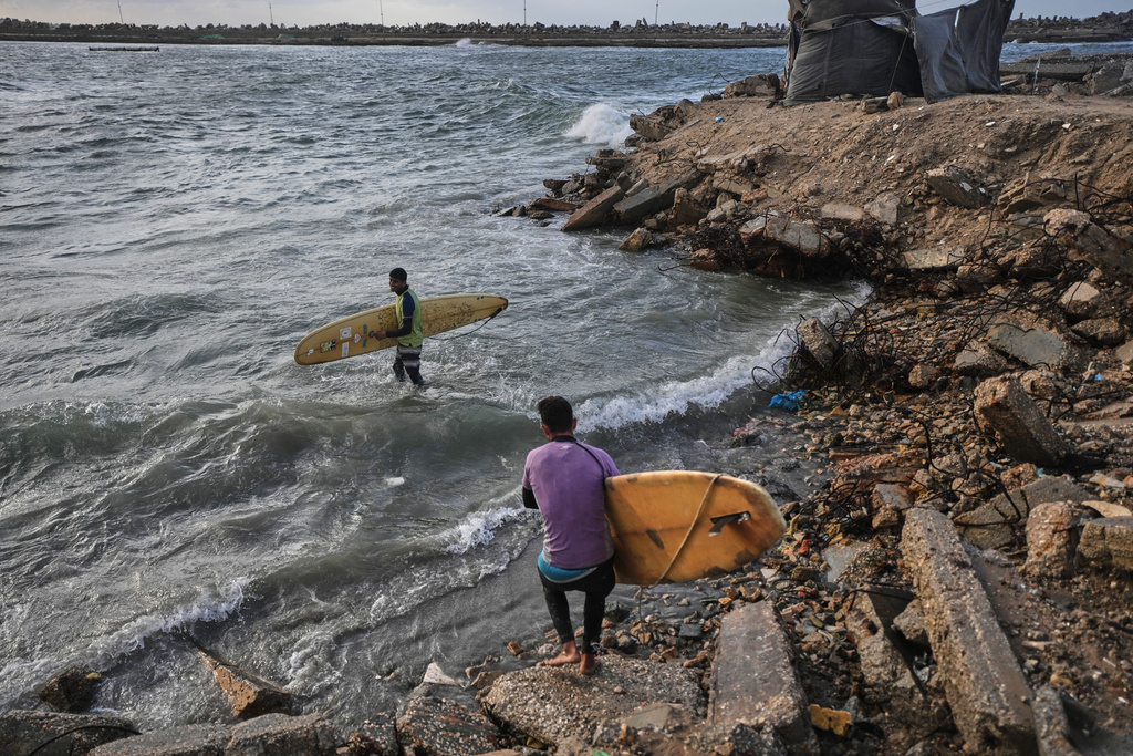 Palestinian Khalil Abu Jayyab, front, and Tahseen Abu Assi carry their boards as they enter the water along a damaged shoreline in Gaza City, Sunday, Dec. 28, 2025. (AP Photo/Jehad Alshrafi)