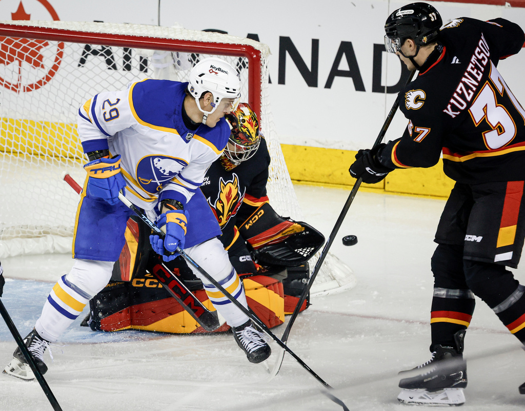 Buffalo Sabres' Beck Malenstyn, left, looks on as Calgary Flames goalie Dustin Wolf deflects a shot while Yan Kuznetsov defends during the first period of an NHL hockey game in Calgary, Alberta, Monday, Dec. 8, 2025. (Jeff McIntosh/The Canadian Press via AP)