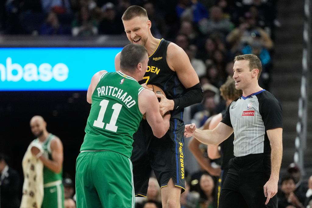 Golden State Warriors center Kristaps Porziņģis, top, smiles while wrestling for the ball with Boston Celtics guard Payton Pritchard (11) as referee Brandon Schwab watches during the second half of an NBA basketball game in San Francisco, Thursday, Feb. 19, 2026. (AP Photo/Jeff Chiu)
