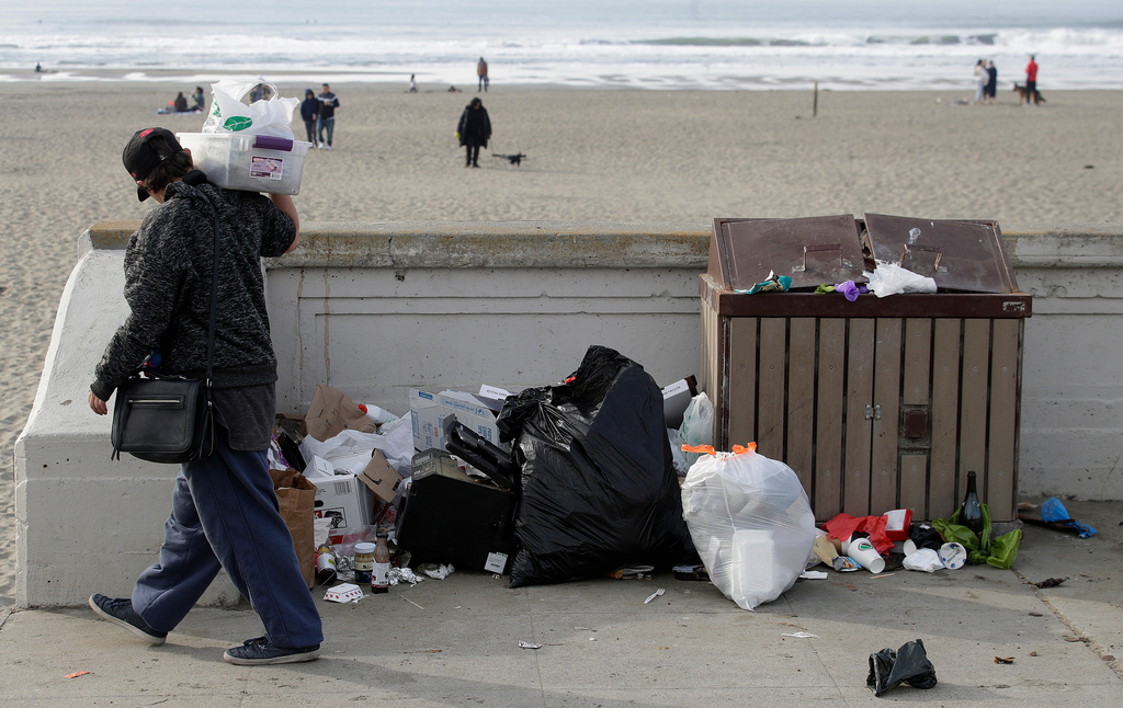 FILE - A woman walks past trash piled next to a garbage bin at Ocean Beach in San Francisco on Jan. 3, 2019. (AP Photo/Jeff Chiu, File)