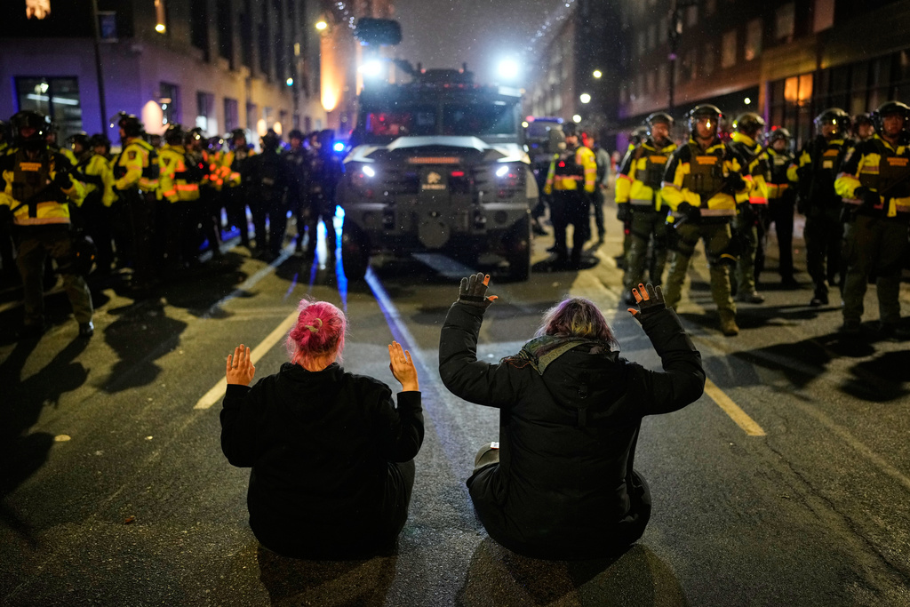 Two people sit in the street with their hands up in front of Minnesota State Patrol during a protest and noise demonstration calling for an end to federal immigration enforcement operations in the city, Friday, Jan. 9, 2026, in Minneapolis. (AP Photo/John Locher)
