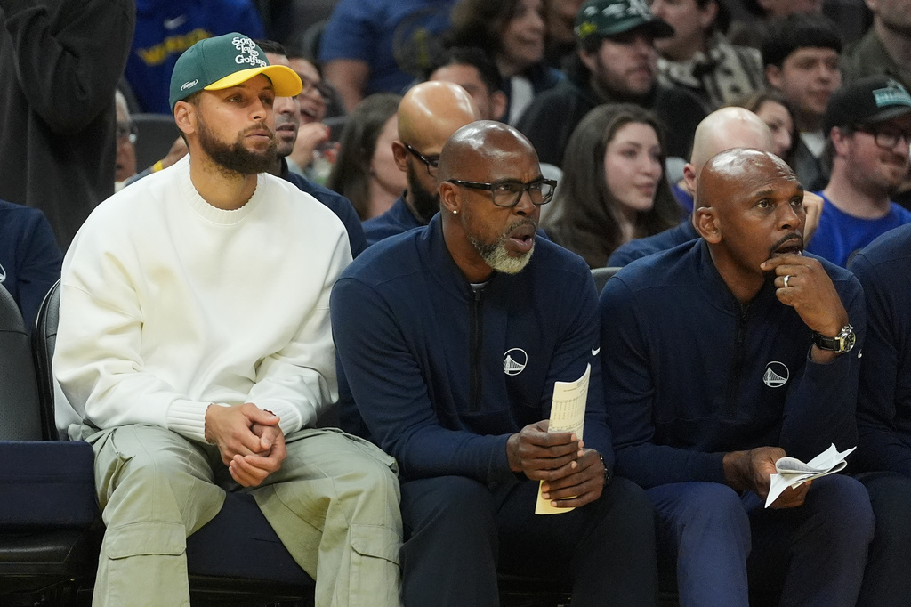 Injured Golden State Warriors guard Stephen Curry, left, watches from the bench with assistant coaches Kris Weems, middle, and Jerry Stackhouse during the first half of an NBA basketball game against the Cleveland Cavaliers in San Francisco, Thursday, April 2, 2026. (AP Photo/Jeff Chiu)