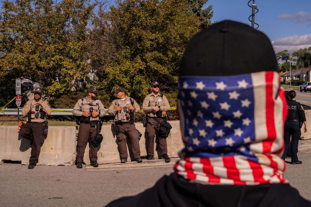 A protestor wearing an American flag face covering stands opposite Cook County Sheriffs outside of an Immigration and Customs Enforcement facility in Broadview, Ill., Saturday, Oct. 11, 2025. (AP Photo/Adam Gray)