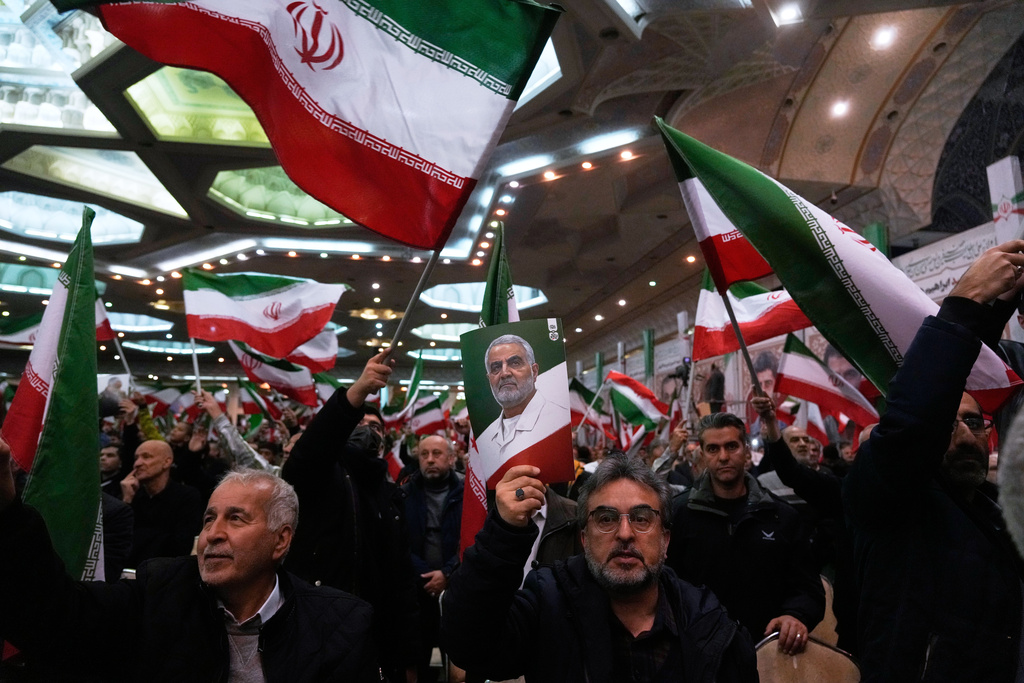 People wave Iranian flags as one of them holds up a poster of the late commander of the Iran's Revolutionary Guard expeditionary Quds Force, Gen. Qassem Soleimani, who was killed in a U.S. drone attack in 2020 in Iraq, during a ceremony commemorating his death anniversary at the Imam Khomeini grand mosque in Tehran, Iran, Thursday, Jan. 1, 2026. (AP Photo/Vahid Salemi)