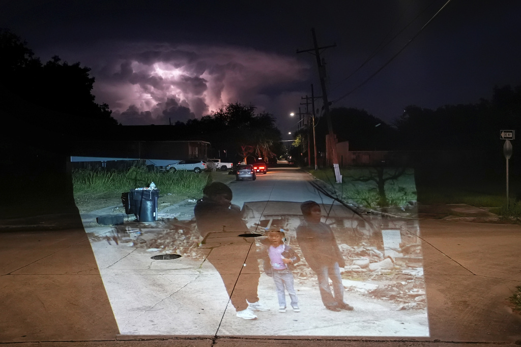 This photo of Valerie Thomas and her nieces Shante Fletcher, 6, and Sarine Fletcher, 11, viewing the destruction of her brother’s home in the aftermath of Hurricane Katrina in the Lower Ninth Ward of New Orleans, taken by AP photographer Gerald Herbert, is projected Wednesday, Aug. 13, 2025, onto the same block, while a heat lightning storm illuminates the clouds. (AP Photo/Gerald Herbert)