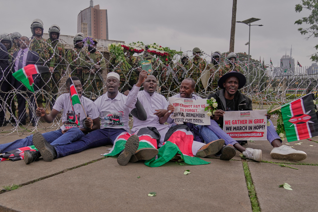Protesters sing and wave Kenyan flags near parliament during a demonstration to mark two years since more than 60 people died in anti-government demonstrations that resulted in the storming of the parliament, as police officers stand behind a razor-wire barricade in Nairobi, Kenya, Thursday, June 25, 2026. (AP Photo/Brian Inganga)