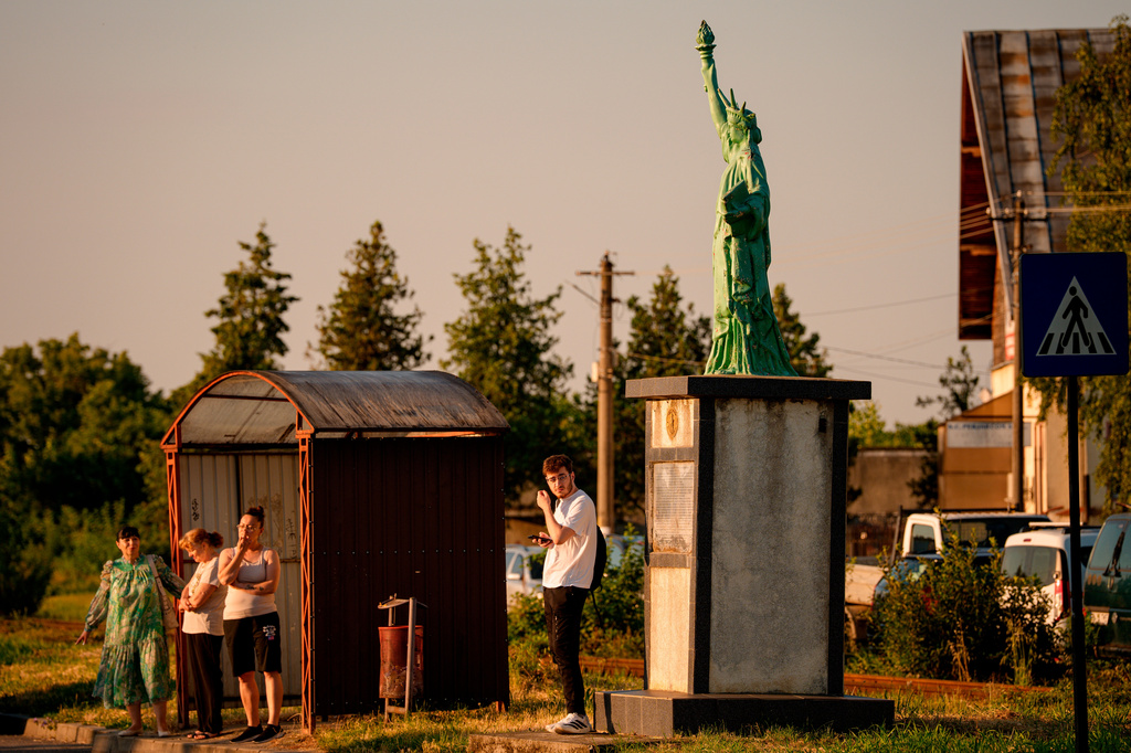 People wait for a bus next to a replica of the Statue of Liberty, made as a symbol of Romanian-American friendship by 90 year old Romanian artist Cristian Petru Balan, who lives in the U.S., in the village of Boldesti–Scaieni in southern Romania, Friday, June 26, 2026. (AP Photo/Vadim Ghirda)