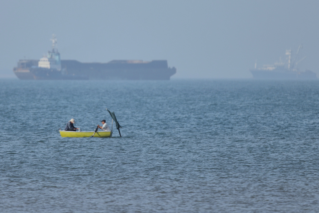 Two men sit in a small boat on the water as cargo ships are anchored in the background in the Strait of Hormuz off Bandar Abbas, Iran, Monday, May 4, 2026. (Amirhosein Khorgooi/ISNA via AP)