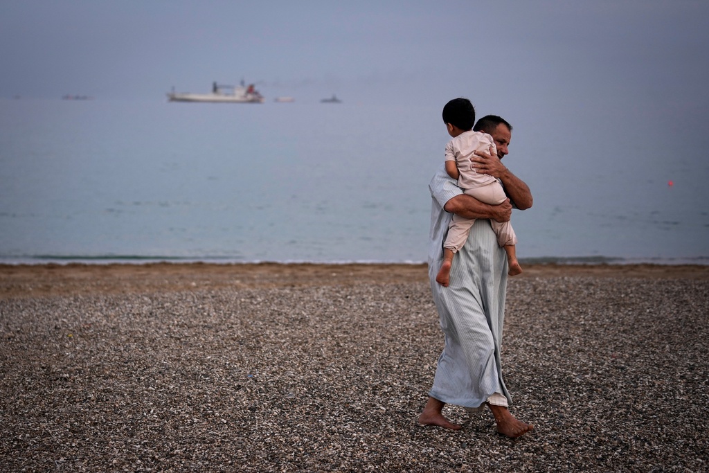 A man carries a child on the beach as oil tankers and cargo ships line up in the Strait of Hormuz near Khor Fakkan, United Arab Emirates, Wednesday, March 11, 2026. (AP Photo/Altaf Qadri)