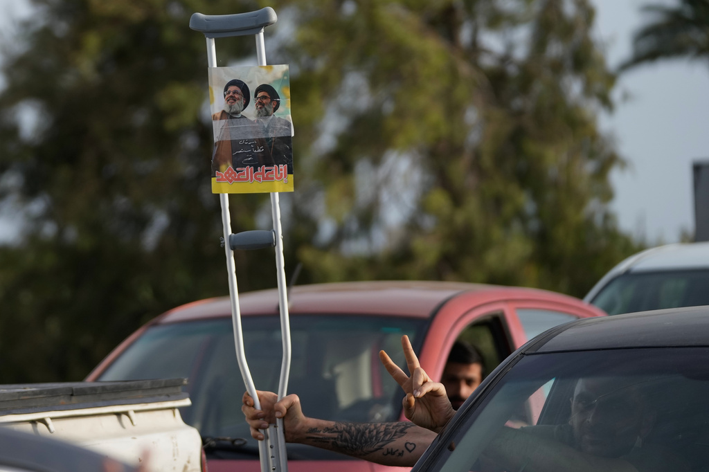 A displaced man flashes victory sign and holds a crutch with a poster showing late Hezbollah leaders Sayyed Hassan Nasrallah and Hashem Safieddine, with writing that reads "on the pledge, we remain" as he returns to his village following a ceasefire between Hezbollah and Israel, in Qasmiyeh near the city of Tyre, south Lebanon, Friday, April 17, 2026. (AP Photo/Mohammed Zaatari)