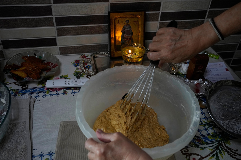 Maria Chanioti, 66, prepares Fanouropita, an orange-flavored cake, as a small icon of Saint Fanourios, known in Greek as "The Revealer," is seen in the background in Athens, Wednesday, Aug. 26, 2026. (AP Photo/Thanassis Stavrakis)