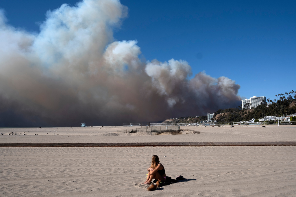 FILE - A lone sunbather sits and watches a large plume of smoke from a wildfire rise over the Pacific Palisades, in Santa Monica, Calif., Jan. 7, 2025. (AP Photo/Richard Vogel, File)