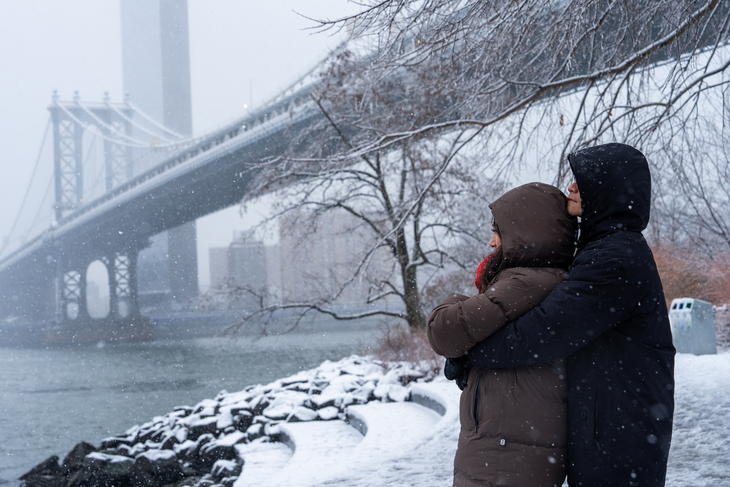 A couple embrace during a snow in front of the Manhattan Bridge, Sunday, Dec. 14, 2025, in the Brooklyn borough of New York. (AP Photo/Adam Gray)