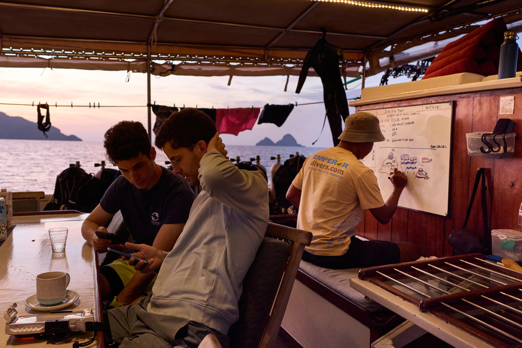 Scuba divers Alejandro Sarró, left, and Manel Sangrà check their phones as dive guide Kristanto Umbu Kudu prepares a briefing aboard the Raja Laut dive boat in Raja Ampat, Indonesia, Tuesday, March 3, 2026. (AP Photo/Claudia Rosel)