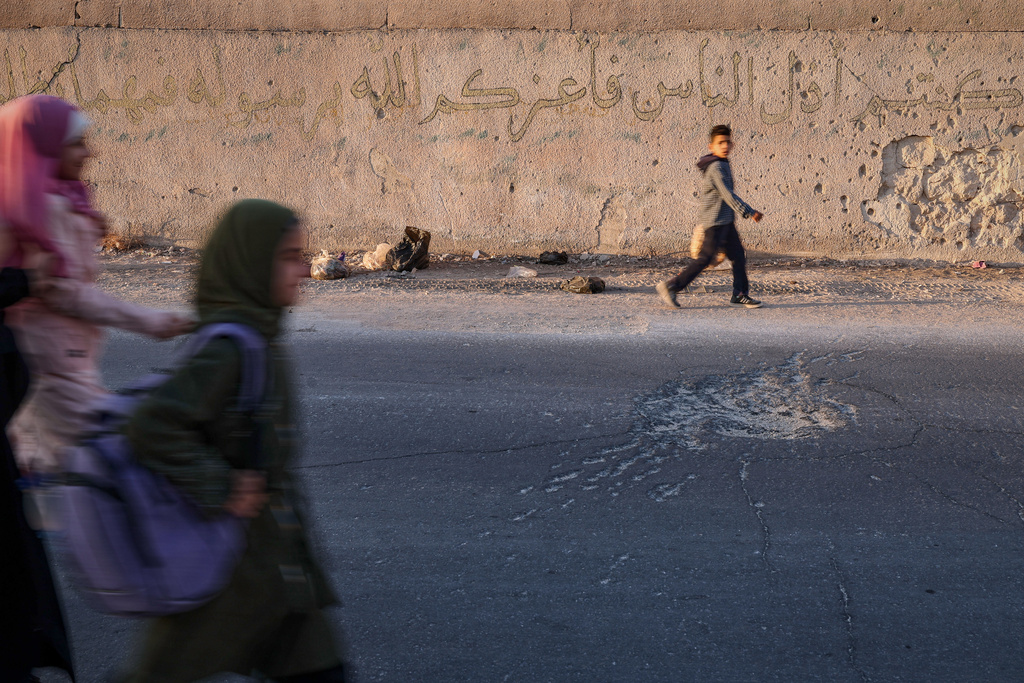 Students walk past a wall riddled with bullet holes and the impact mark of a mortar shell on the street, dating back to the fighting between forces loyal to former President Bashar Assad and rebel groups, in the village of Maar Shmarin, in the Idlib countryside, Syria, Sunday, Oct. 19, 2025. (AP Photo/Ghaith Alsayed)