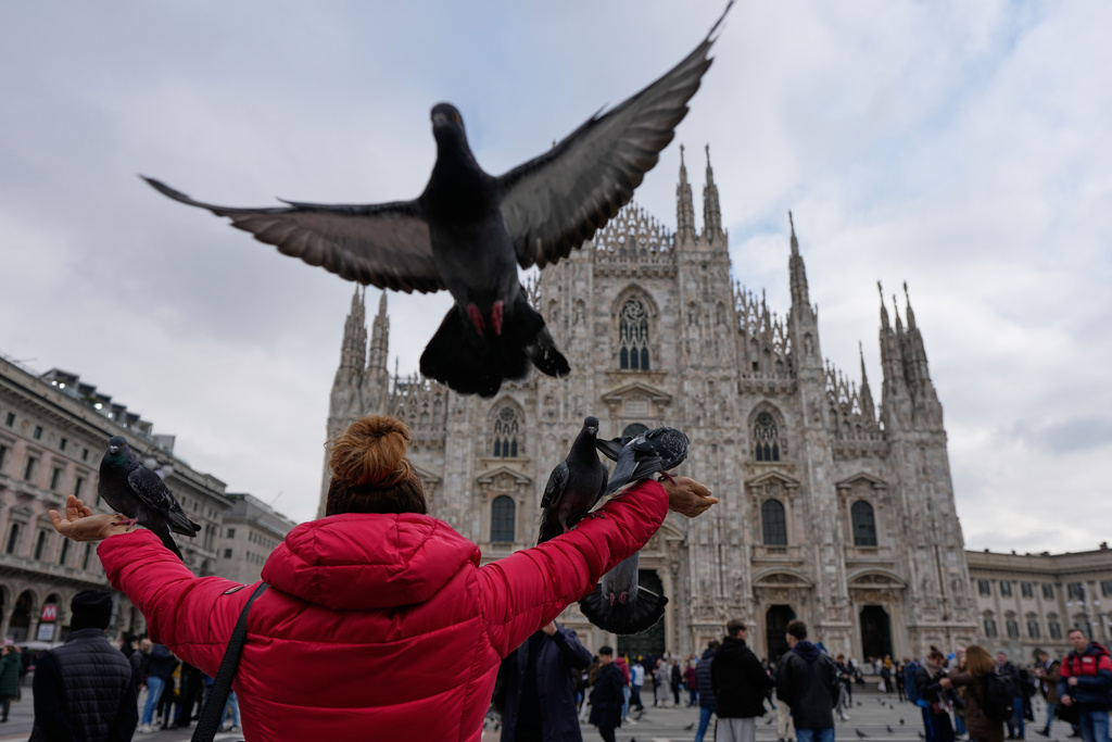 A woman feeds pigeons as they gather around her in Piazza del Duomo, with the Duomo cathedral in the background, during the 2026 Winter Olympics in Milan, Italy, Wednesday, Feb. 11, 2026. (AP Photo/Hassan Ammar)