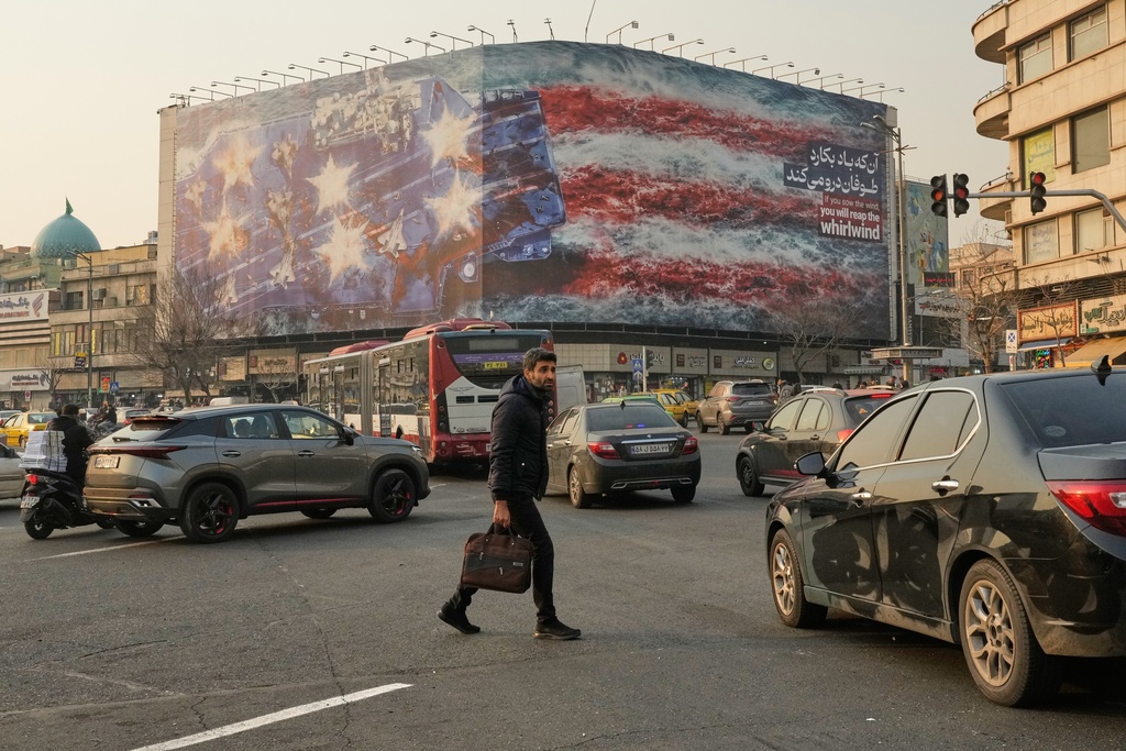 A man walks among vehicles in front of a billboard depicting a damaged U.S. aircraft carrier with disabled fighter jets on its deck and a sign reading in Farsi and English, "If you sow the wind, you'll reap the whirlwind," at Enqelab-e-Eslami (Islamic Revolution) Square in Tehran, Iran, Sunday, Jan. 25, 2026. (AP Photo/Vahid Salemi)
