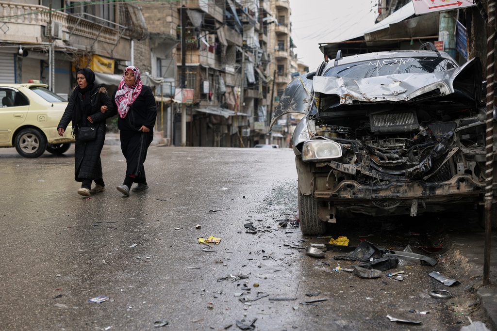 Women walk by a damaged car in the predominantly Kurdish Sheikh Maqsoud neighborhood after days of fighting between government forces and Kurdish fighters in the northern city of Aleppo, Syria, Tuesday, Jan. 13, 2026. (AP Photo/Ghaith Alsayed)