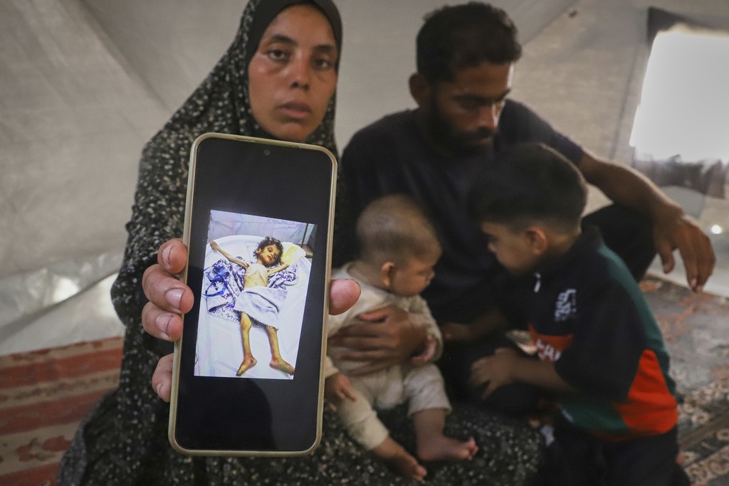 Sitting next to her husband, Amin, and two of their children, Fatma Mashi shows a photo on her cellphone of the body of her 2½-year-old daughter, Ro'a, who doctors say had no preexisting conditions and died of severe malnourishment, at their tent in Muwasi, southern Gaza Strip, Friday, Aug. 8, 2025. Experts say it's not unusual for one family member to fare worse than the others. (AP Photo/Mariam Dagga)