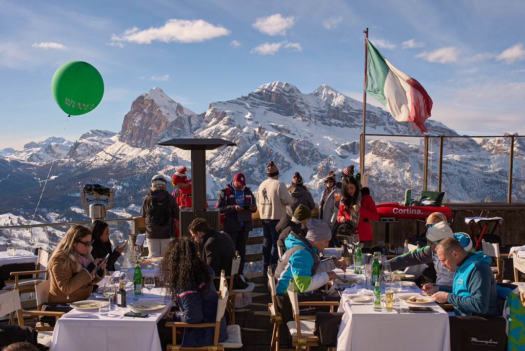 People enjoy a beverage on Faloria Mountain, with a scenic view of the Olympic alpine ski runs behind them, during the 2026 Winter Olympics, in Cortina d'Ampezzo, Italy, Friday, Feb. 13, 2026. (AP Photo/Jacquelyn Martin)