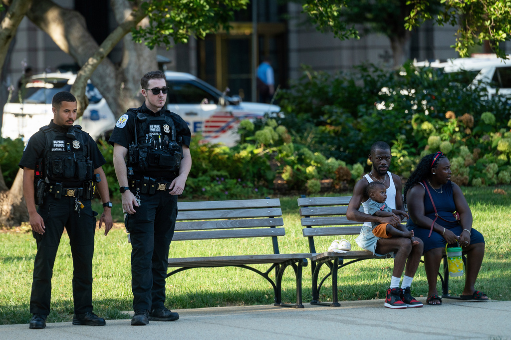 A family sits on a bench in Franklin Park in downtown Washington as Washington Metropolitan Police patrol Thursday, Aug. 14, 2025. (AP Photo/Cliff Owen)