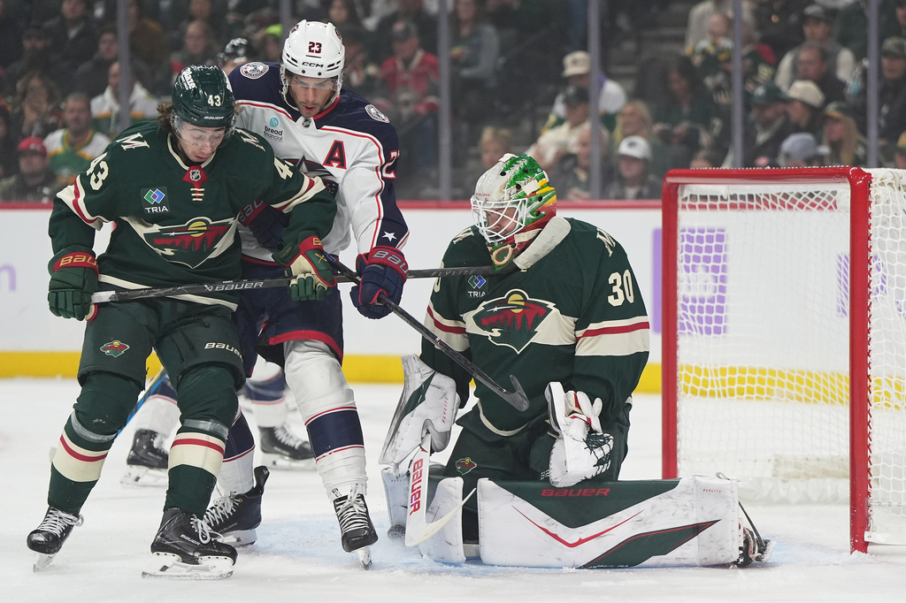 Minnesota Wild goaltender Jesper Wallstedt (30) and defenseman Quinn Hughes (43) defend against a shot as Columbus Blue Jackets center Sean Monahan (23) pressures them during the first period of an NHL hockey game, Sunday, Oct. 18, 2026, in St. Paul, Minn. (AP Photo/Abbie Parr)