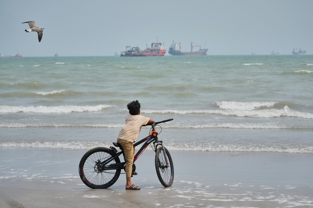 A gull flies overhead as a boy stands with his bicycle on a beach overlooking commercial vessels anchored in the Strait of Hormuz off Bandar Abbas, Iran, Monday, Sept. 7, 2026. (AP Photo/Vahid Salemi)