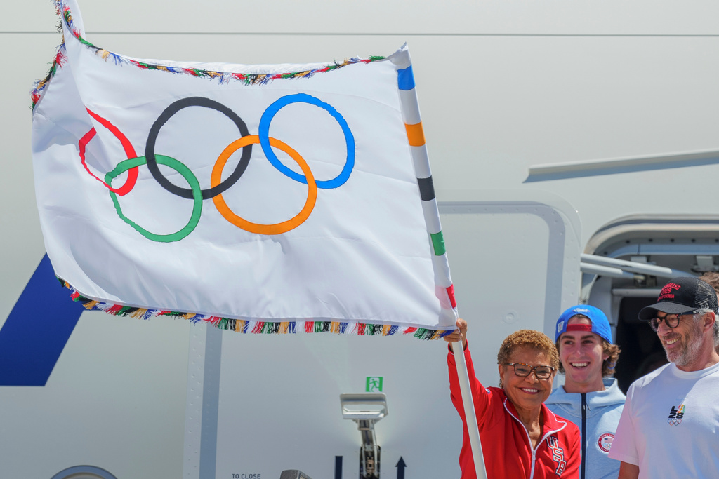 FILE - Los Angeles Mayor Karen Bass, left, holds the official Olympic flag, next to LA28 chairman Casey Wasserman, right, and Team USA Oympian skateboarder Tate Carew, center, at Los Angeles International Airport on Aug. 12, 2024. (AP Photo/Damian Dovarganes, File)