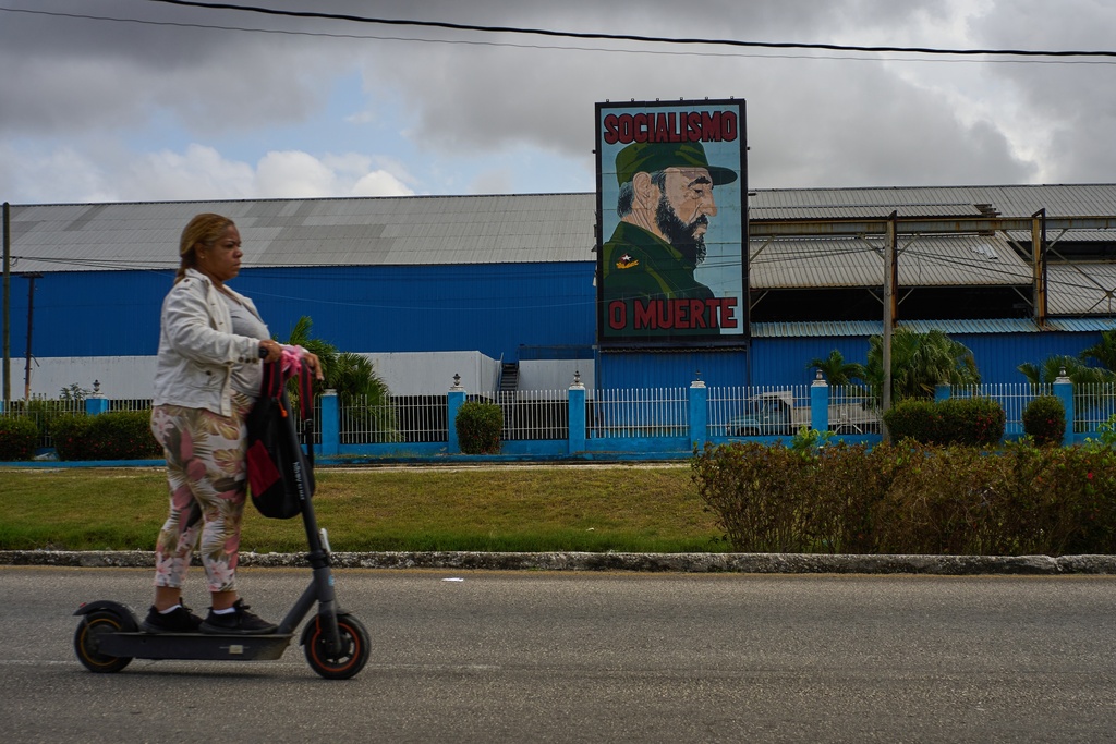 A woman rides an electric scooter past a factory displaying an image depicting the late Cuban leader Fidel Castro, bearing the words "Socialism or Death", in Havana, Cuba, Thursday, March 19, 2026. (AP Photo/Ramon Espinosa)