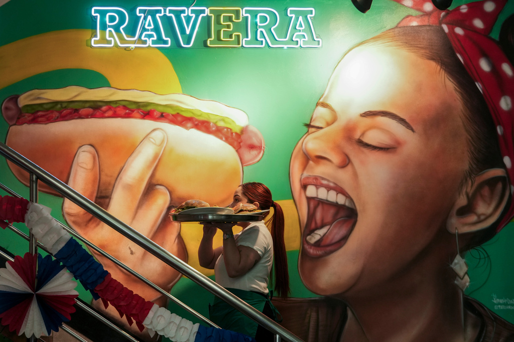 A waitress carries a tray with "completos", traditional Chilean hot dogs topped with mashed avocado, diced tomatoes and mayonnaise, past a mural dedicated to the dish at the Ravera restaurant in downtown Santiago, Chile, Thursday, Sept. 10, 2026. (AP Photo/Esteban Felix)