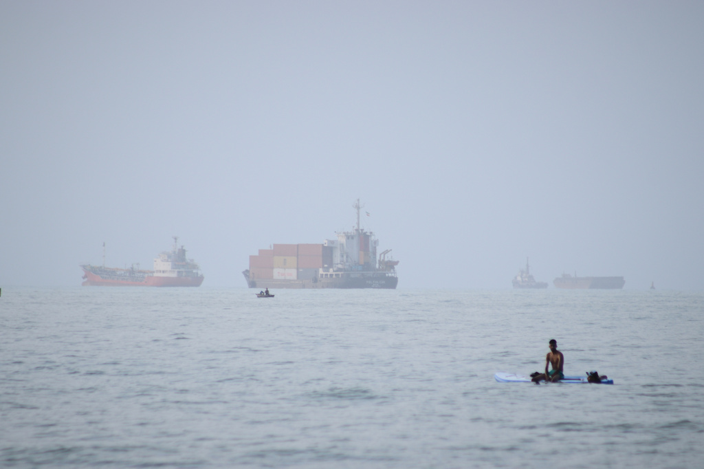 A man sits on a surfboard as a container ship and other commercial vessels appear anchored in the Strait of Hormuz off Bandar Abbas, Iran, Monday, July 27, 2026. (Razieh Poudat/ISNA via AP)