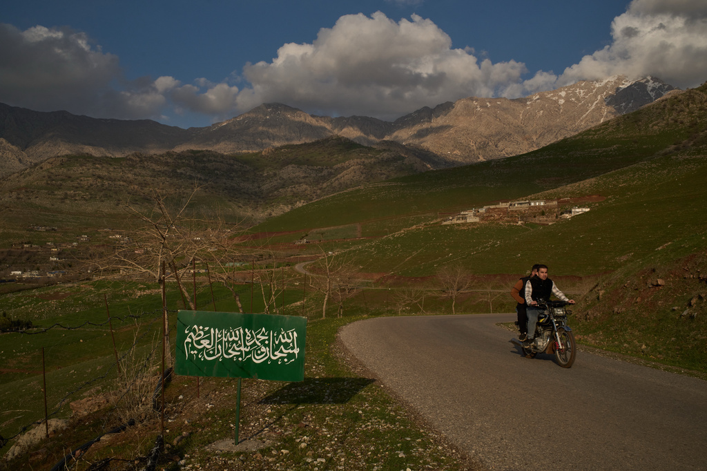A man rides his motorcycle past a sign bearing a prayer in Arabic reading "Glory and praise be to Allah; glory be to Allah, the Great," along a road near the Iraq-Iran border in the mountainous Kurdish region near Halabja, Iraq, Tuesday, March 17, 2026. (AP Photo/Leo Correa)