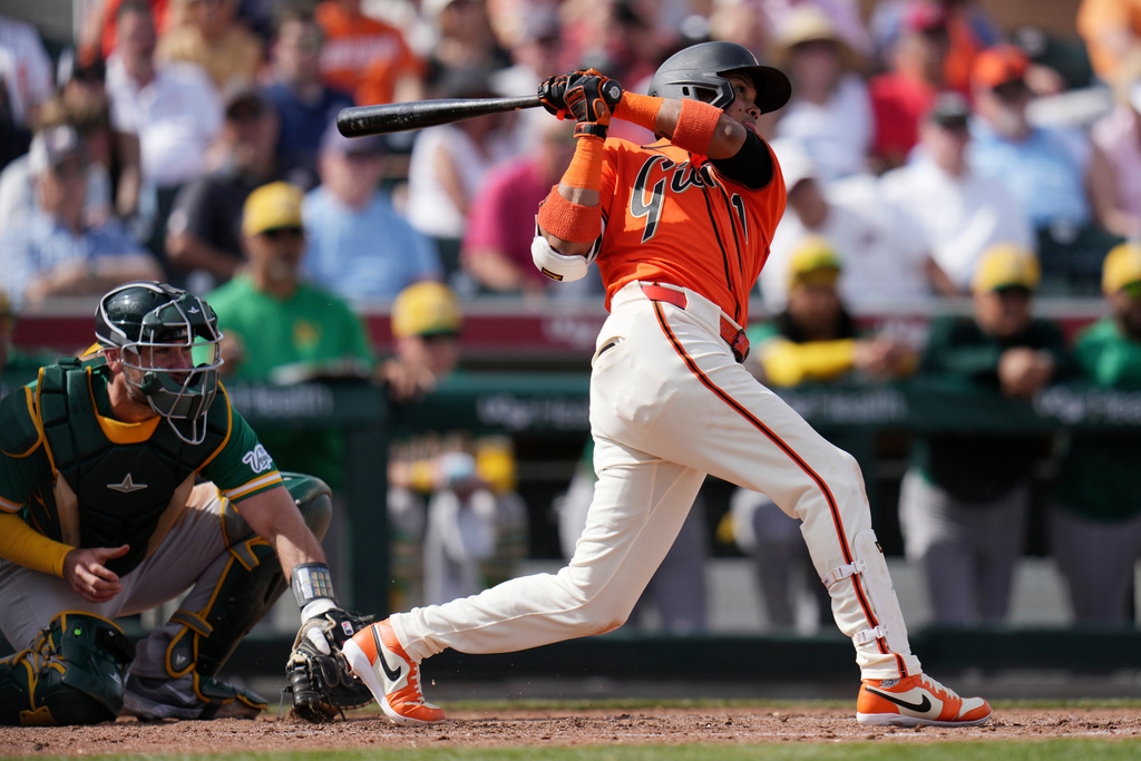 San Francisco Giants' Luis Arraez, right, connects for a run-scoring single as Athletics catcher Austin Wynns, left, looks on during the third inning of a spring training baseball game Monday, Feb. 23, 2026, in Scottsdale, Ariz. (AP Photo/Ross D. Franklin)
