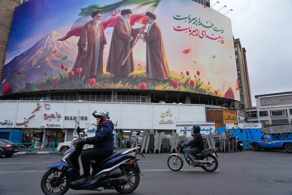 FILE - Motorbikes drive past a billboard depicting Iran's late Supreme Leader Ayatollah Ali Khamenei, center, handing the country's flag to his son and successor, Ayatollah Mojtaba Khamenei, right, as the late revolutionary founder Ayatollah Ruhollah Khomeini stands at left, in a square in downtown Tehran, Iran, March 10, 2026. (AP Photo/Vahid Salemi, File)