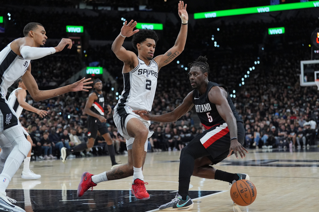 Portland Trail Blazers guard Jrue Holiday (5) drives against San Antonio Spurs guard Dylan Harper (2) and San Antonio Spurs forward Victor Wembanyama, left, during the first half in Game 2 of a first-round NBA playoffs basketball series in San Antonio, Tuesday, April 21, 2026. (AP Photo/Eric Gay)