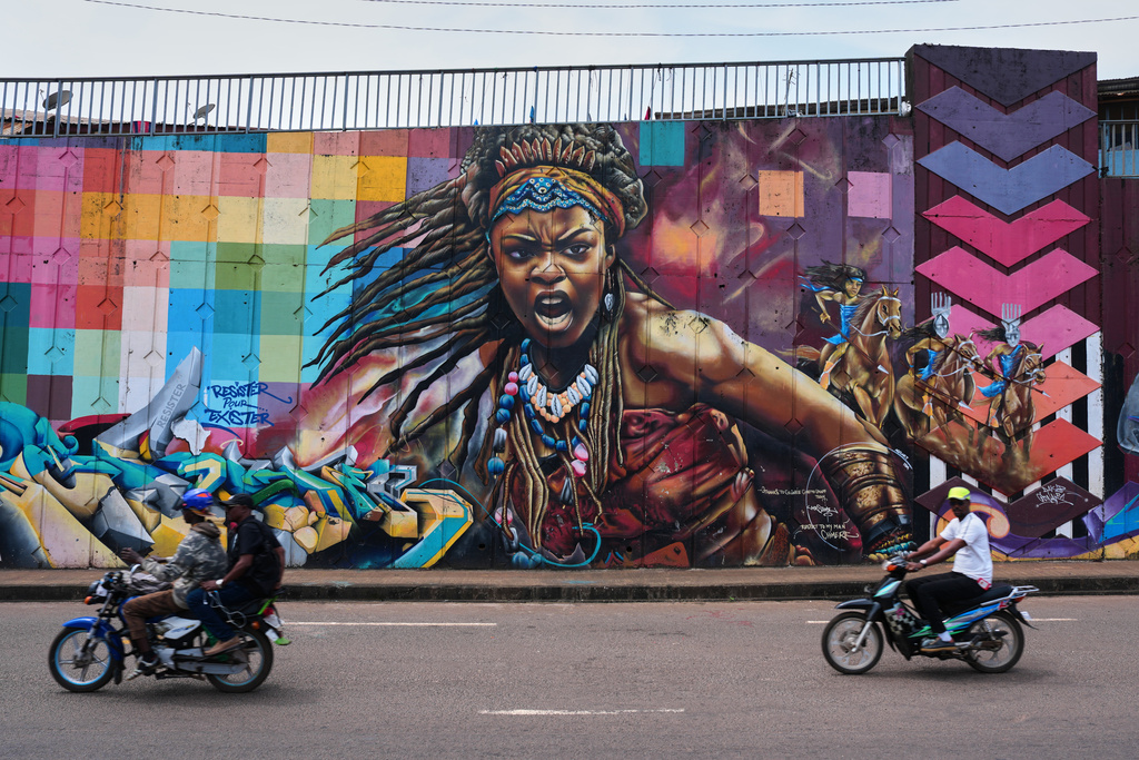 Motorcyclists ride past a mural depicting a powerful woman on a street wall in Conakry, Guinea, Sunday, Oct. 19, 2025. (AP Photo/Misper Apawu)