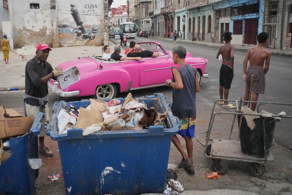 A man collects trash while tourists ride around in a classic American car during a power outage in Havana, Cuba, Wednesday, Dec. 3, 2025. (AP Photo/Ramon Espinosa)