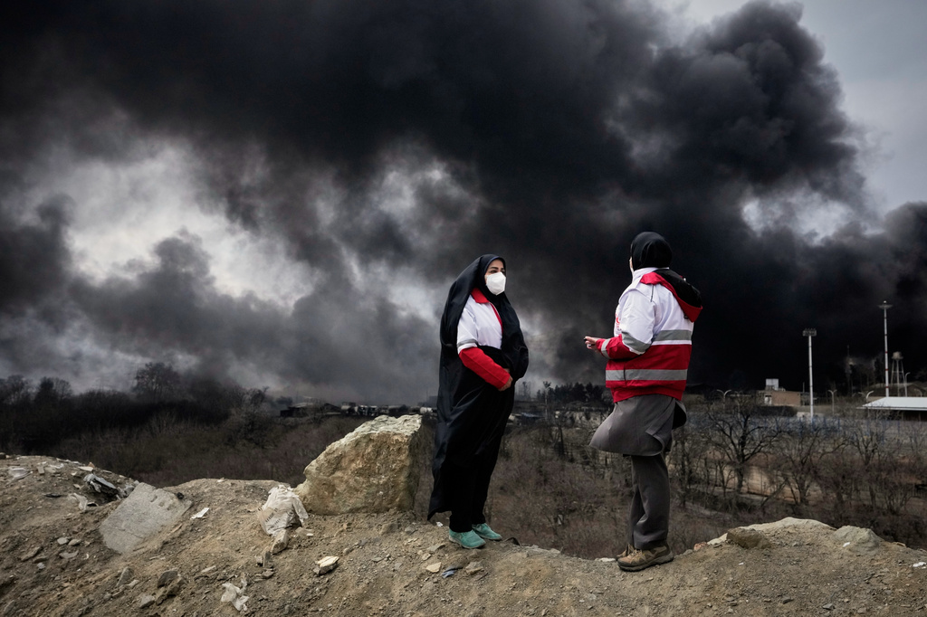 FILE - Two women from the Iranian Red Crescent Society stand as a thick plume of smoke from a U.S.-Israeli strike on an oil storage facility late Saturday rises into the sky in Tehran, Iran, Sunday, March 8, 2026. (AP Photo/Vahid Salemi, File)