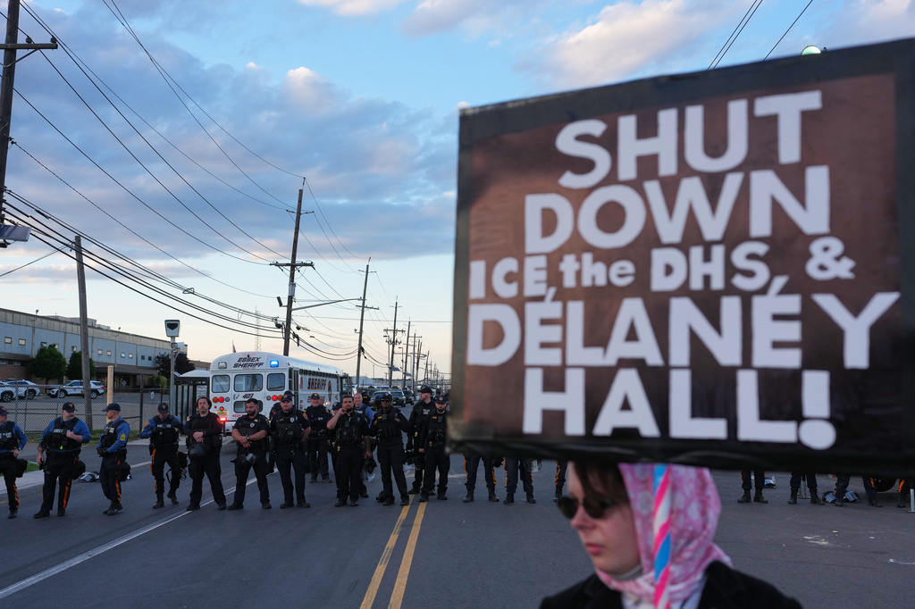A protester holds a sign as law enforcement officers stand outside Delaney Hall detention center Sunday, May 31, 2026, in Newark, N.J. (AP Photo/Adam Gray)