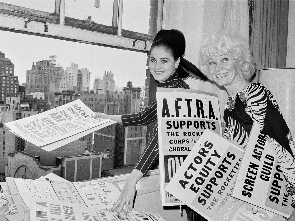 FILE — Rockettes Lynda Farmer, left, and Penny Singleton, pose like they're ready to toss picket signs out the window of the American Guild of Variety Artists headquarters, at the Hotel Warwick, Oct. 12, 1967, in New York, following a general settlement of a contract dispute between Radio City Music Hall and the Rockettes and other performers. (AP Photo/Eddie Adams, File)