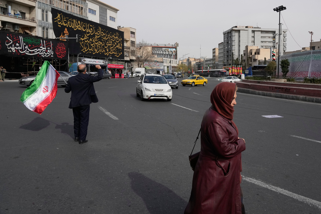 A man waves an Iranian flag during a campaign in support of the government as a woman and vehicles pass by at the Enqelab-e-Eslami, or Islamic Revolution, square in downtown Tehran, Iran, Sunday, March 22, 2026. (AP Photo/Vahid Salemi)
