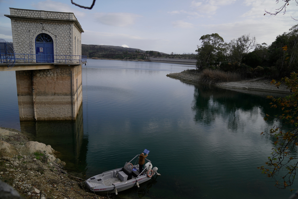 Giorgos Chrysinas Water Board (EYDAP) electricity supervisor adjusts a Greek flag on a small boat at Lake Marathon near Athens, Greece, on Tuesday, Jan. 6, 2026, as receding water levels reflect years of low rainfall. (AP Photo/Thanassis Stavrakis)