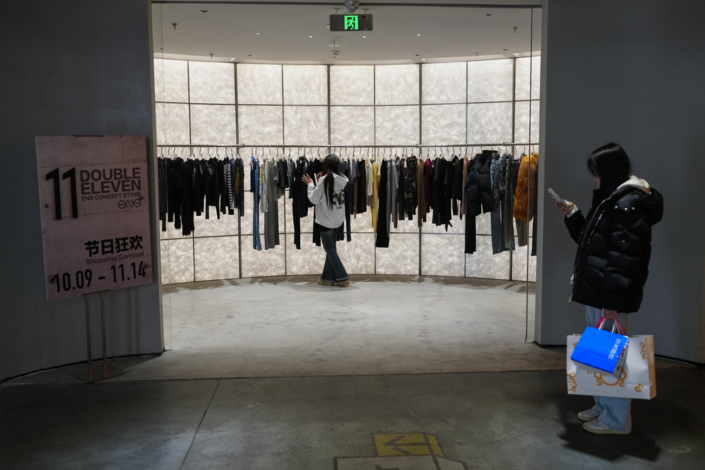 A woman with her purchased goods browses a smartphone as a worker arranges clothing at a fashion boutique store displaying a poster to promote Singles' Day discounts at a shopping mall in Beijing on Nov. 10, 2025. (AP Photo/Andy Wong)